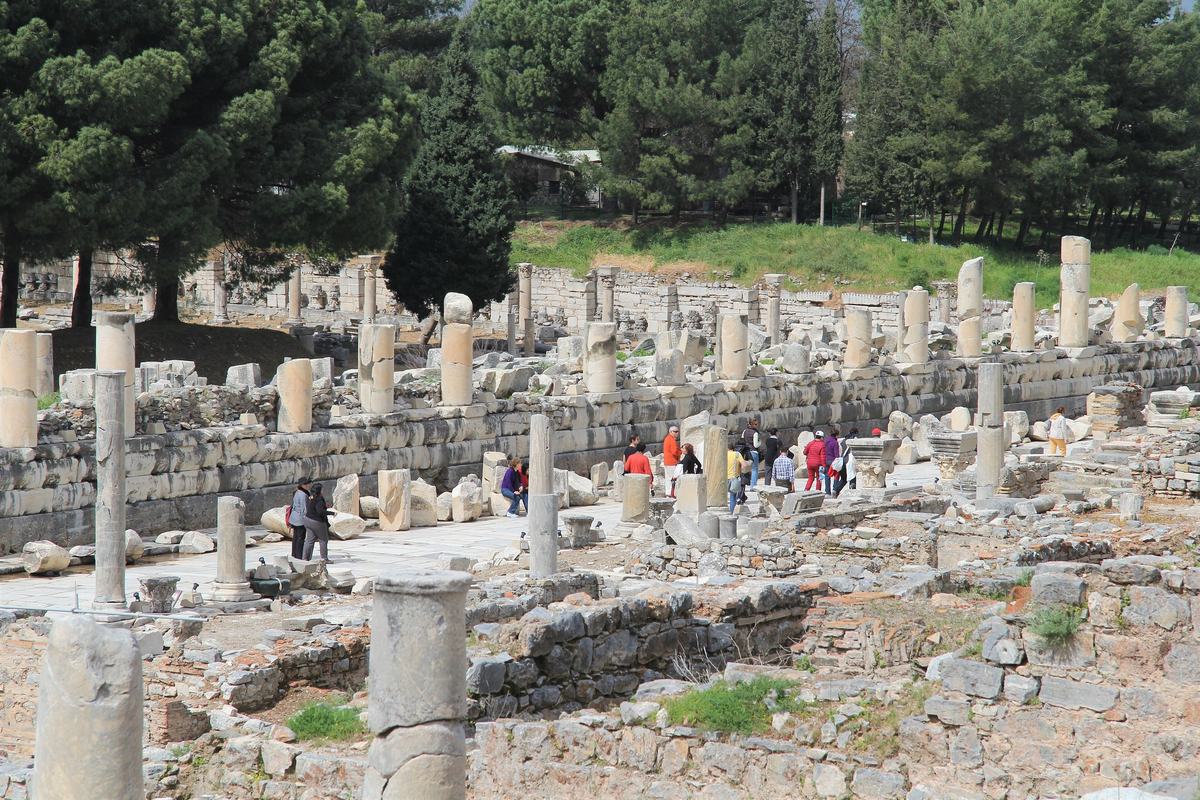 Pequeños grupos de visitantes recorren la calle de Mármol de Éfeso junto al muro del ágora, con pinos al fondo, en un día tranquilo de primavera