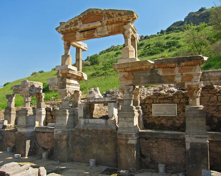 Restos de la fuente de Trajano en Éfeso, con columnas y un arquitrabe remontados sobre el frente de la fuente y la ladera verde detrás