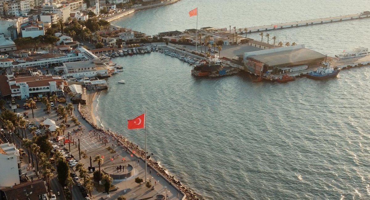 Vista aérea del paseo marítimo de Kuşadası al atardecer, con una gran bandera turca en la plaza junto al mar y, al fondo, los muelles del puerto de cruceros
