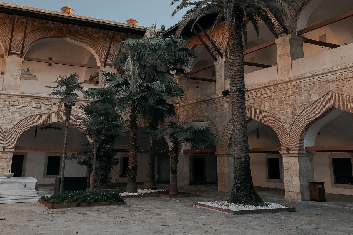 Patio del caravasar de Öküz Mehmed Paşa en Kuşadası, con arcadas de piedra y ladrillo en dos pisos, palmeras y una fuente de mármol