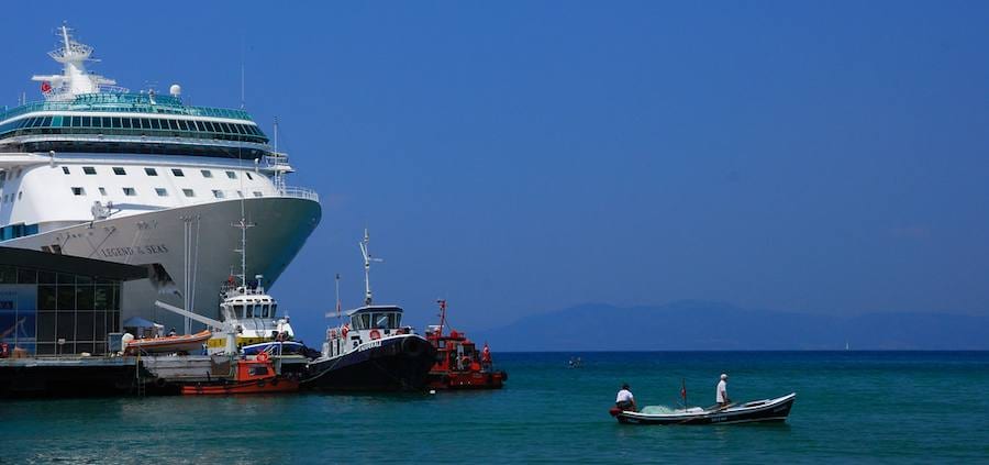 Un crucero atracado en la terminal de Kuşadası, con remolcadores bajo la proa y una barca de pesca faenando en el agua turquesa de delante
