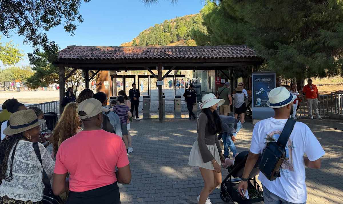 Visitantes caminando hacia los tornos de la Puerta Sur de Éfeso, bajo un tejadillo de teja, con pinos y la ladera del monte Bülbül al fondo